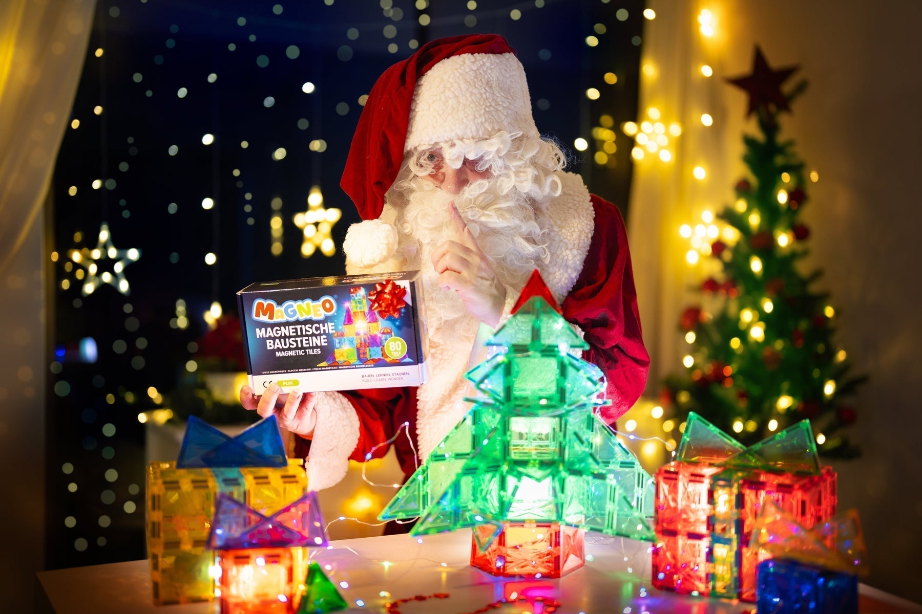 Weihnachtsszene mit einem verkleideten Weihnachtsmann, der die Magneo 80-Teile Box hält, umgeben von magnetischen Baustein-Bauten wie einem leuchtenden Weihnachtsbaum und Geschenkboxen.